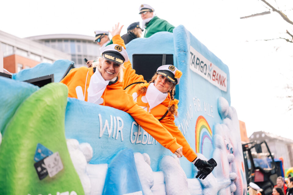 Eine Gruppe von Personen in orangefarbenen Kostümen auf einem festlich dekorierten Wagen. Im Hintergrund ist eine blaue Wand mit dem Logo der TARGOBANK und dem Text "WIR GEBEN ALLES" sichtbar. Die Personen tragen Hüte und posieren fröhlich.