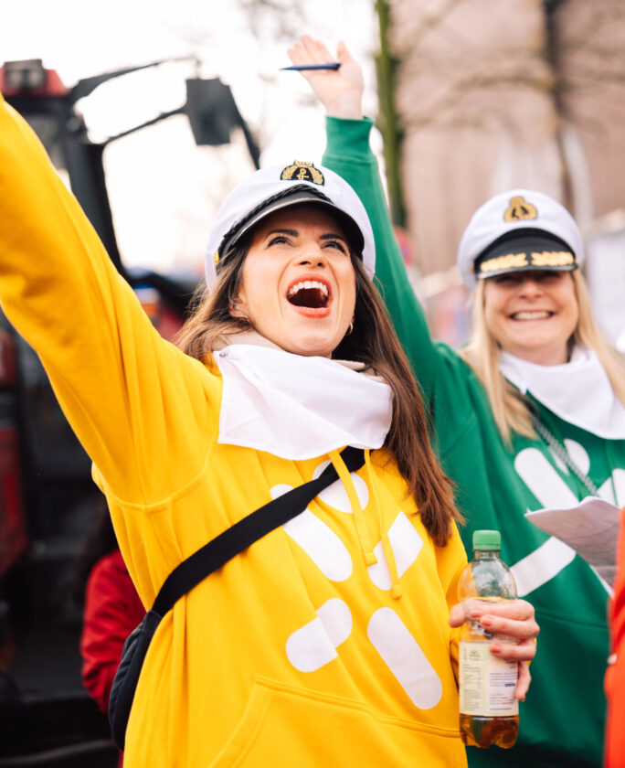 Zwei Personen stehen auf einer Veranstaltung. Eine trägt einen gelben Pullover mit weißen Mustern und hält eine Flasche in der Hand. Die andere trägt einen grünen Pullover. Beide haben Kapitänsmützen auf und scheinen fröhlich zu sein.