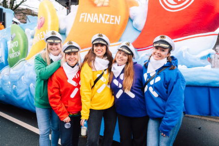 Fünf Personen stehen vor einem bunten Wagen, der mit Wolken und Symbolen dekoriert ist. Sie tragen Kapitänsmützen und farbige Pullover: rot, gelb, grün und blau. Jede hat ein weißes Tuch um den Hals. Im Hintergrund sind große bunte Plakate mit den Worten 'FINANZEN' und anderen Begriffen sichtbar.