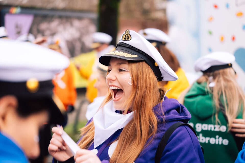 Eine Gruppe von Personen in festlichen Outfits mit Schiffsmützen. Eine Person mit langen, roten Haaren trägt einen lila Pullover und hält ein Papier in der Hand. Im Hintergrund sind weitere Personen in verschiedenen bunten Kleidungen zu sehen, darunter eine grüne Jacke mit dem Text "die ganze breite".