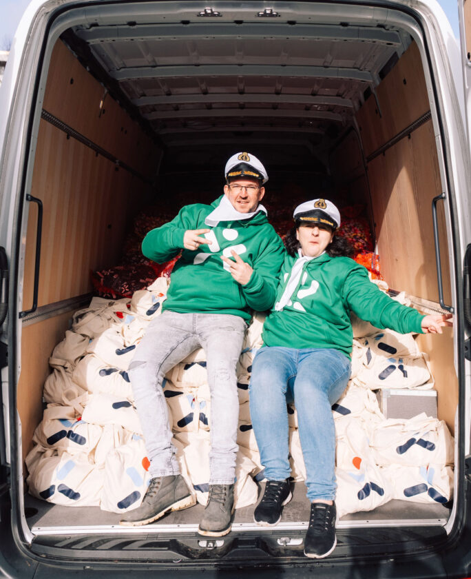 Zwei Personen sitzen in einem Lieferwagen, der mit weißen Taschen gefüllt ist. Beide tragen grüne Sweatshirts und Kapitänsmützen. Die Taschen sind ordentlich gestapelt, und die Atmosphäre wirkt fröhlich.