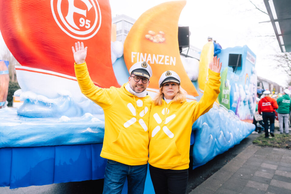 Ein Paar in gelben Kapuzenpullovern posiert vor einem bunten Festwagen mit dem Schriftzug 'FINANZEN'. Im Hintergrund sind weitere Teilnehmer in bunten Outfits zu sehen.