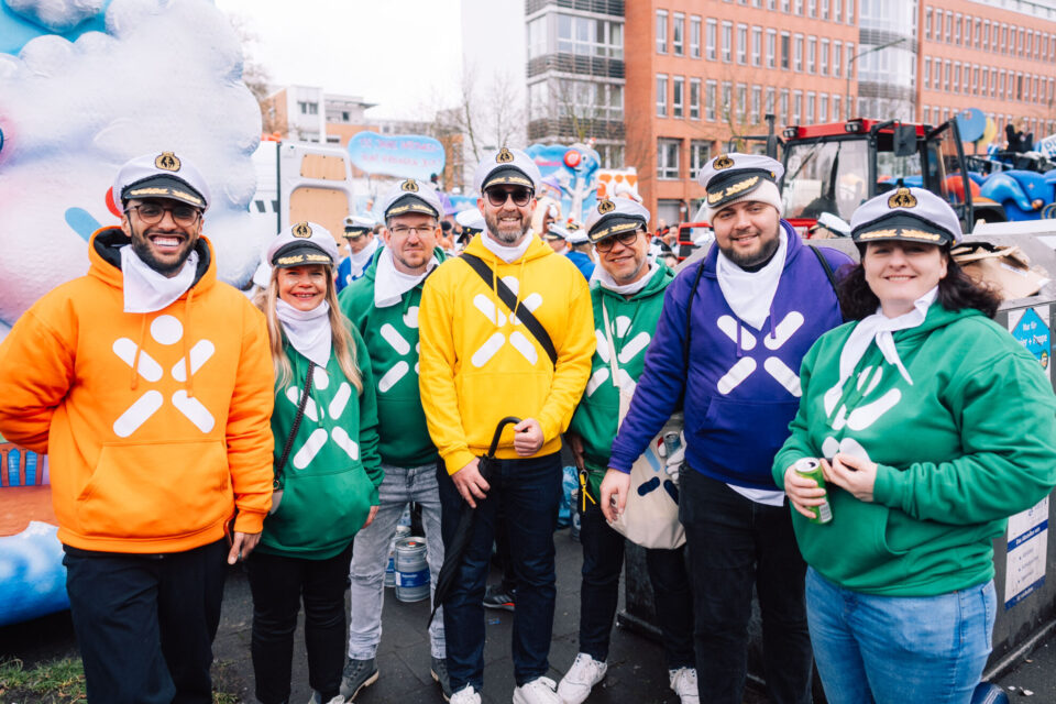 Eine Gruppe von sieben Personen steht in bunten Hoodies (orange, grün, gelb, lila) und weißen Halstüchern. Sie tragen Mützen mit einem Logo. Im Hintergrund sind festliche Elemente und Gebäude sichtbar.