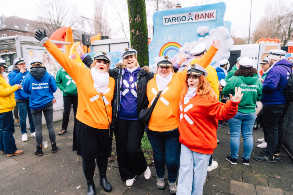Eine Gruppe von Menschen in bunten Kapuzenpullovern posiert fröhlich. Einige tragen Hüte. Im Hintergrund ist ein Werbebanner der TARGO Bank zu sehen, das "WIND FÜR ALLE" und einen Regenbogen zeigt. Die Atmosphäre wirkt festlich.