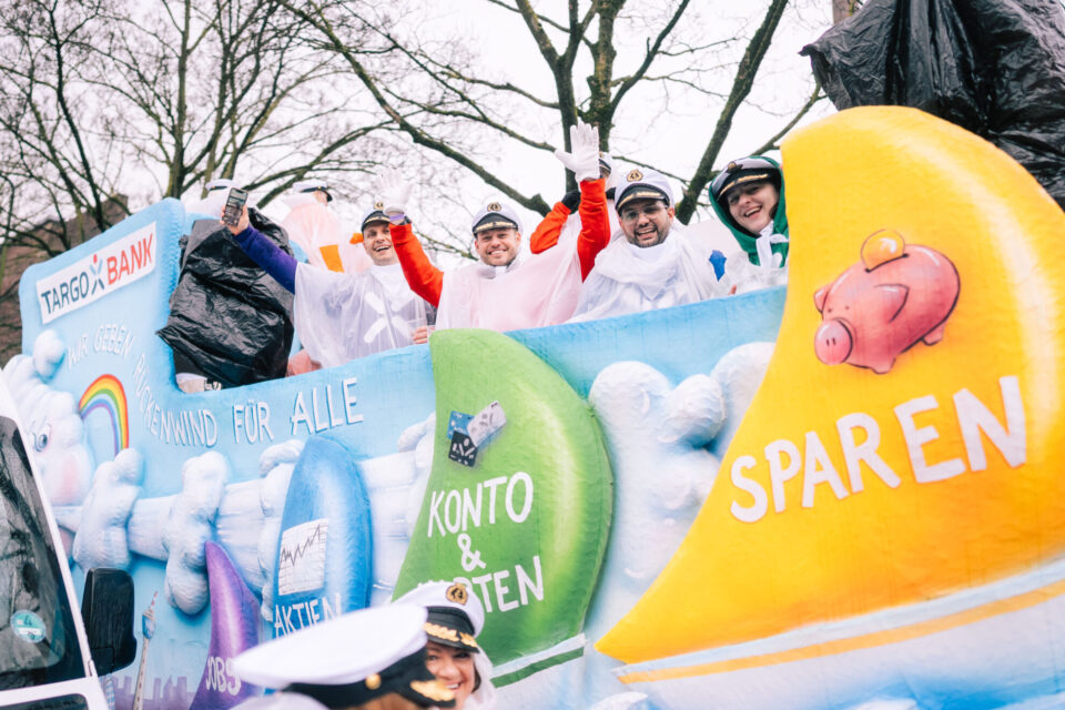 Eine Gruppe von Menschen steht auf einem bunten Festwagen, der mit Wolken und verschiedenen Symbolen bemalt ist. Sie tragen weiße Regenmäntel und Hüte. Der Wagen zeigt die Aufschrift 'TARGOBANK' und andere Texte wie 'KONTO & KARTEN' und 'SPAREN'. Im Hintergrund sind Bäume und ein grauer Himmel zu sehen.