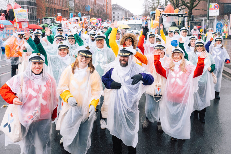 Eine Gruppe von Menschen in durchsichtigen Regenponchos steht auf einer Straße. Sie winken und haben unterschiedliche farbige T-Shirts darunter. Einige tragen Caps. Im Hintergrund sind festliche Umzugswagen und eine Menschenmenge sichtbar.
