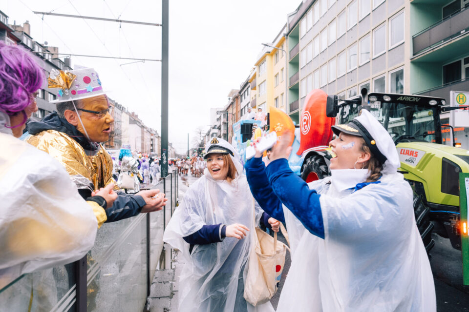 Eine bunte Karnevalsfeier auf der Straße mit mehreren Menschen in Kostümen. Einige tragen Regenponchos, während andere festliche Hüte tragen. Im Hintergrund sieht man eine Parade mit bunten Wagen und Menschen in verschiedenen Kostümen. Ein Mann hält etwas in der Hand, während eine andere Person ihm etwas entgegenhält. Ein Traktor steht in der Nähe auf der Straße.