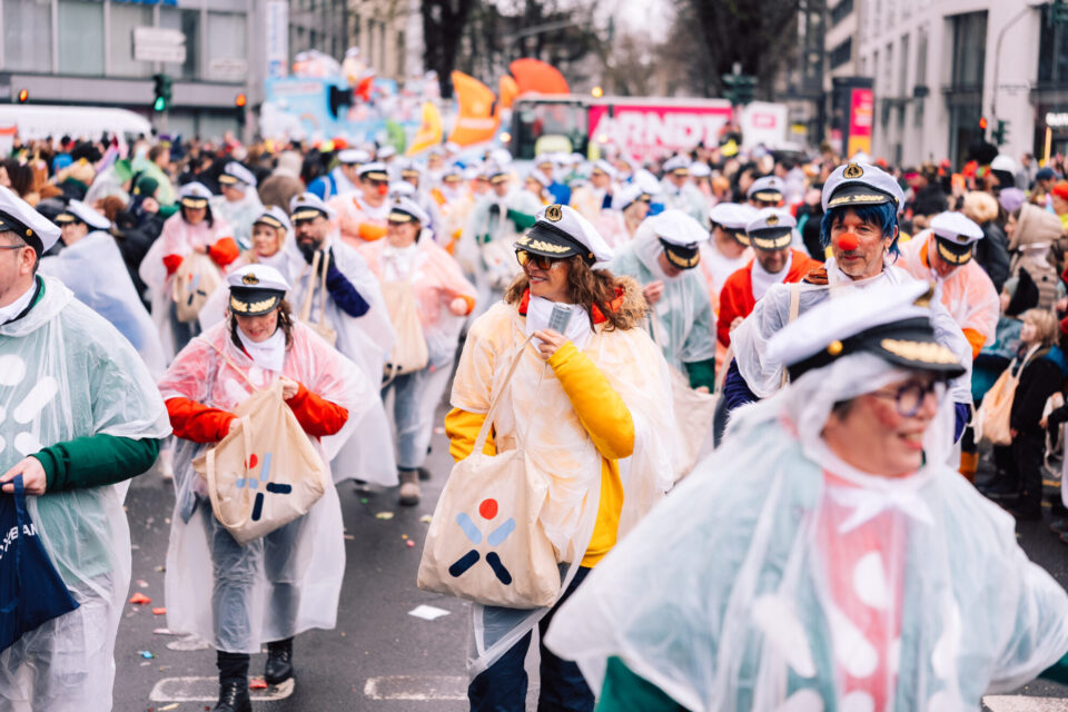 Eine Menschenmenge nimmt an einem Umzug teil, während sie Regenponchos trägt. Viele Personen tragen Kapitänsmützen und halten Taschen. Die Szene zeigt einen Straßenumzug mit bunten Kostümen und Plakaten im Hintergrund.