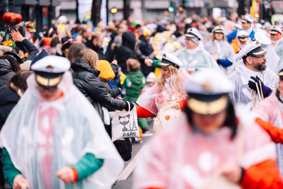 Eine belebte Straße während eines Karnevals, mit vielen Menschen in Regenmänteln. Einige tragen Hüte und Accessoires, während andere Taschen halten. Die Menge ist farbenfroh und lebhaft.