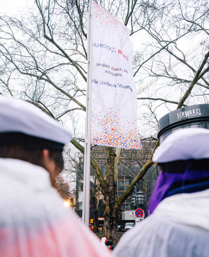Die Flagge mit dem Logo von ARGO und BNY hängt an einem Baum. Auf der Flagge steht: 'Jeder Job ist anders, Wir lieben Vielfalt.' Im Vordergrund sind Personen mit weißen Hüten und bunten Umhängen zu sehen.