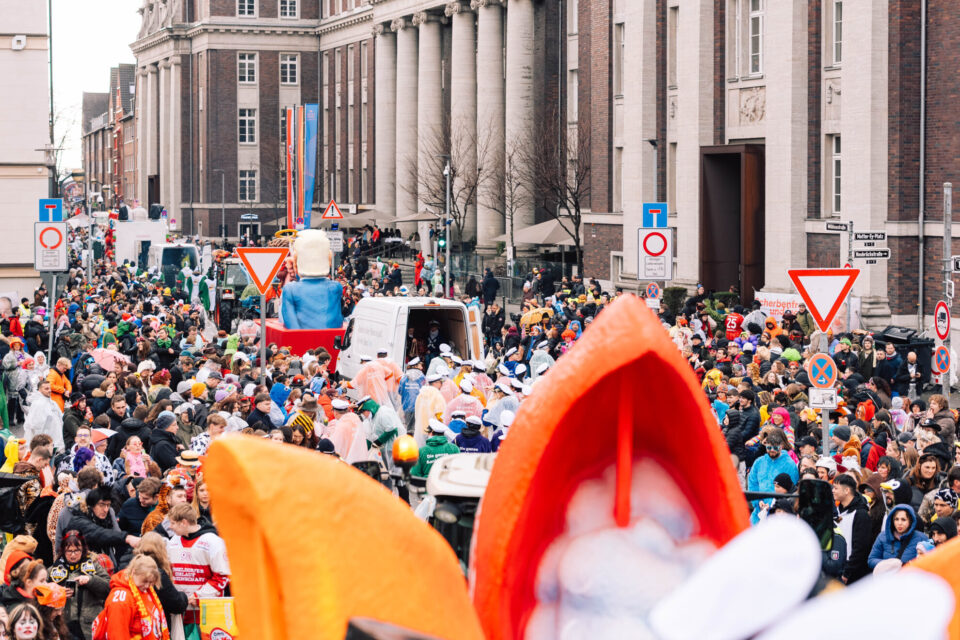 Eine große Menschenmenge versammelt sich auf einer Straße während einer Veranstaltung, mit bunten Kostümen und Festwagen im Hintergrund. Verkehrsschilder sind sichtbar, und einige Menschen tragen Regenponchos. Im Hintergrund sind Gebäude mit Säulen und Schildern zu sehen.