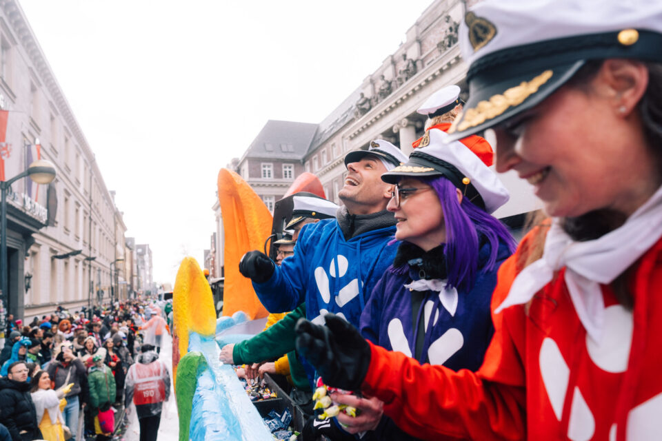 Eine Gruppe von Menschen in bunten Kostümen steht auf einem Wagen während einer Parade. Sie tragen Hüte und zeigen ihre Hände. Im Hintergrund sind Menschenmengen auf der Straße, die die Feierlichkeiten beobachten. Gebäude sind entlang der Straße sichtbar.
