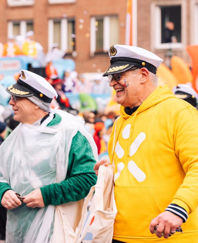 Zwei Personen in einem Festumzug, eine trägt ein grünes Sweatshirt und eine durchsichtige Regenjacke, die andere ein gelbes Sweatshirt mit einem weißen Logo. Beide halten eine Tasche und wirken amüsiert vor einer bunten Menschenmenge und einer festlichen Kulisse.