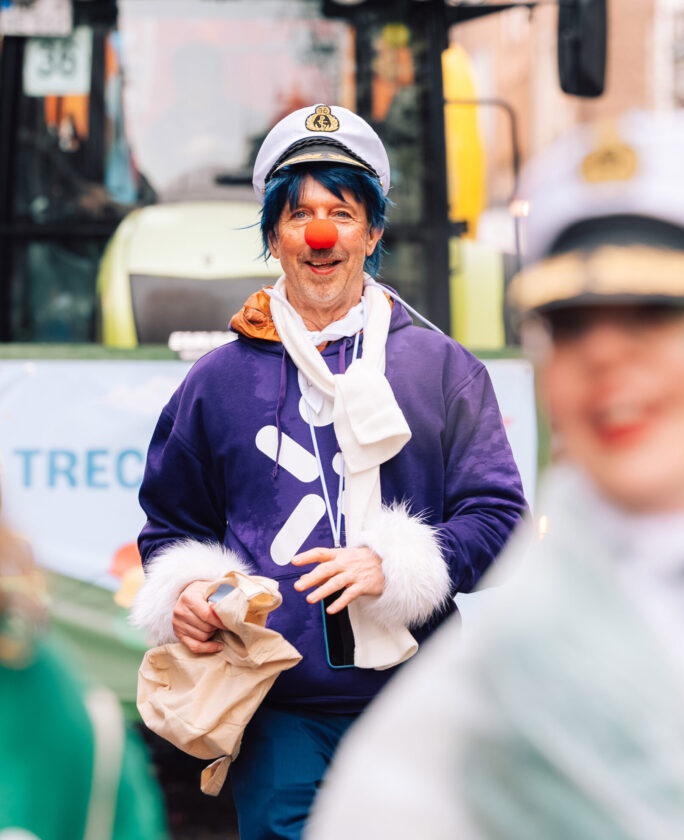Eine Person in einem lila Sweatshirt mit einem weißen Symbol und einem weißen Schal. Sie trägt eine Schirmmütze und hält eine braune Tüte. Im Hintergrund ist ein Fahrzeug und ein Banner sichtbar.