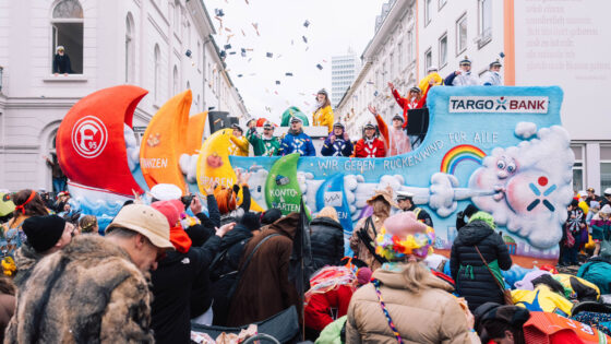 Eine bunte Parade mit Menschen in festlichen Kostümen, die auf einem großen Wagen stehen. Der Wagen hat eine farbenfrohe Gestaltung mit Symbolen wie Wellen und Wolken. Auf dem Banner steht "WIR GEBEN RÜCKENWIND FÜR ALLE" und das Logo der TARGOBANK ist sichtbar. Im Hintergrund sind Menschen in der Menge, die jubeln und feiern. Über der Parade fliegen Konfetti und kleine Gegenstände.