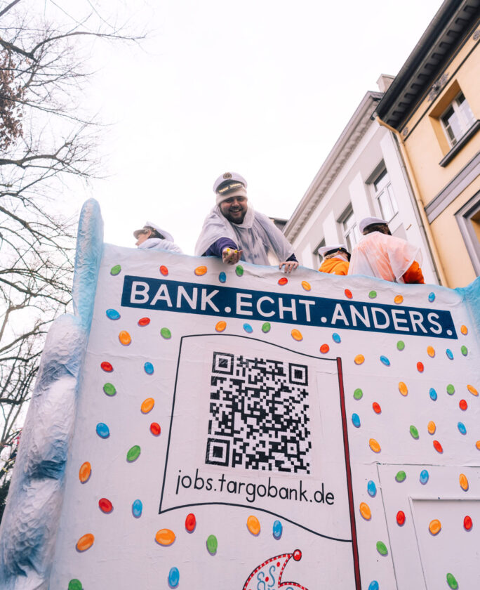 Ein bunter Umzugswagen mit dem Slogan 'BANK.ECHT.ANDERS.' ist zu sehen. Auf der Seite des Wagens befindet sich ein QR-Code und die Website jobs.targobank.de. Der Wagen ist mit bunten, runden Süßigkeiten verziert, und Personen in Schutzkleidung sind oben auf dem Wagen.
