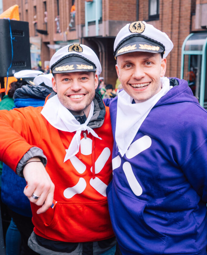 Zwei Personen stehen zusammen auf einer Veranstaltung. Die Person links trägt einen roten Kapuzenpullover mit einem weißen Logo und hat ein weißes Halstuch. Die Person rechts trägt einen lila Kapuzenpullover, ebenfalls mit einem weißen Logo, und hat ein weißes Halstuch. Im Hintergrund sind Menschen und Gebäude zu sehen.