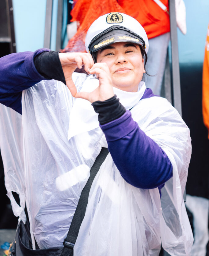 Eine Person in einem weißen Poncho und einer Schirmmütze zeigt mit beiden Händen ein Herzzeichen. Im Hintergrund sind unscharfe Gegenstände und Menschen zu sehen.