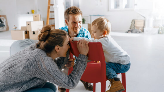 Eine Familie sitzt in einem hellen Raum. Eine Person beugt sich über einen kleinen roten Stuhl, während ein Kind darauf sitzt. Die Erwachsene und das Kind scheinen sich zu unterhalten oder zu spielen. Im Hintergrund sind Kisten und eine Leiter sichtbar, die auf eine Umzugs- oder Renovierungsumgebung hindeuten.