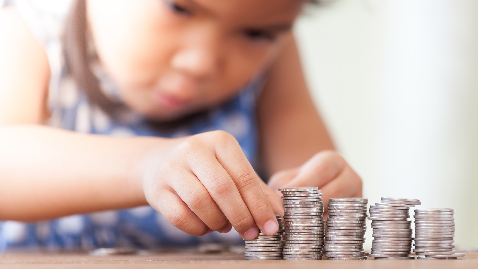 Ein Kind zählt Münzen auf einem Tisch. Die Hände sind sichtbar, während sie Münzen in gestapelter Form anordnen. Es gibt mehrere Stapel unterschiedlich hoher Münzen, die sorgfältig platziert sind.