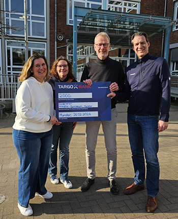 Vier Personen stehen im Freien vor einem Gebäude. Eine Person hält einen großen Scheck von TARGOBANK über 2000 Euro. Die anderen Personen stehen neben ihr und lächeln.