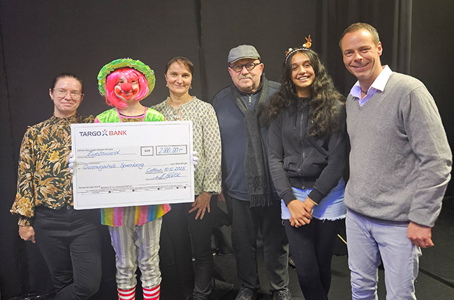 Eine Gruppe von fünf Personen steht vor einem schwarzen Hintergrund. Eine Person in einem bunten Clownskostüm hält einen großen Scheck der TARGO BANK, dessen Betrag 2.000,00 € beträgt. Die anderen Personen sind in unterschiedlichen Outfits gekleidet. Die Gesichter der Menschen sind anonymisiert.