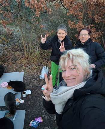 Eine Gruppe von drei Personen steht draußen in der Natur, umgeben von Bäumen und Laub. Im Vordergrund sind mehrere schwarze Katzen und Essen auf einer grauen Unterlage zu sehen. Zwei Personen machen Peace-Zeichen, während eine dritte Person im Hintergrund steht. Alle tragen warme Kleidung.