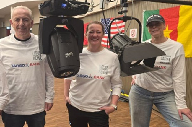 Drei Personen stehen in einem Raum, tragen weiße T-Shirts mit dem Aufdruck 'TARGOBANK'. Eine Kamera ist über ihnen montiert. Im Hintergrund hängen Flaggen, darunter die US-Flagge und die Flagge Kameruns.