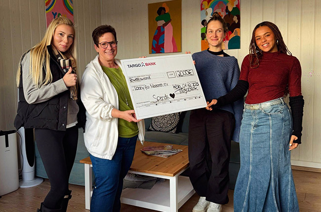 Eine Gruppe von vier Personen steht in einem Raum, in dem eine große Scheckpräsentation stattfindet. Eine Frau in der Mitte hält den Scheck von der TARGO Bank, auf dem eine Spende vermerkt ist. Die anderen drei Personen stehen neben ihr, lächelnd und in legerer Kleidung. Im Hintergrund sind farbenfrohe Kunstwerke an der Wand zu sehen, und Möbel sind im Raum verteilt.