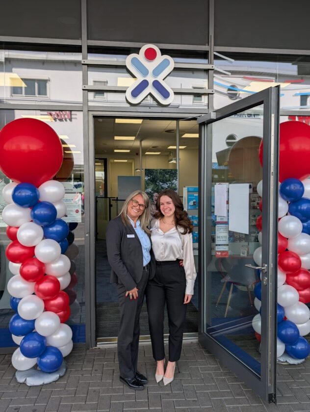 Zwei Frauen stehen vor einem Geschäft, umgeben von bunten Luftballons in Rot, Weiß und Blau. Eine Frau trägt einen schwarzen Anzug und eine blaue Bluse, während die andere eine weiße Bluse und schwarze Hosen trägt. Über ihnen ist ein Logo sichtbar, und die Tür des Geschäfts steht offen.