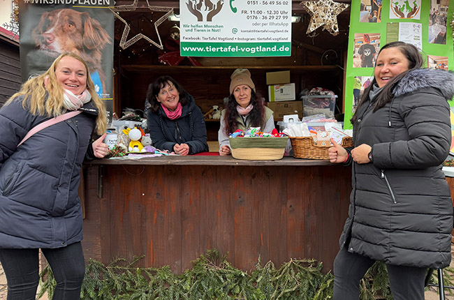 Eine Gruppe von vier Frauen steht an einem Verkaufsstand. Der Stand hat eine Holztheke und ist mit Weihnachtsdekoration geschmückt. Im Hintergrund sind Produkte und Flyer sichtbar. Eine Frau hält einen kleinen Plüschartikel. Die anderen lächeln und interagieren am Stand. Es ist eine kalte Jahreszeit, da die Frauen warme Jacken tragen.