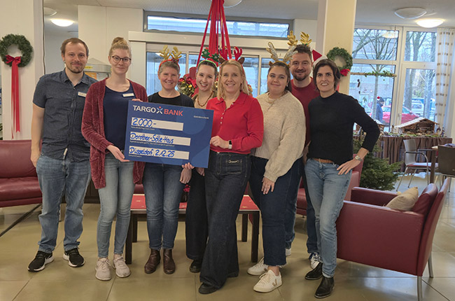 Eine Gruppe von sieben Personen steht in einem Raum, in dem Weihnachtsdekorationen sichtbar sind. Sie halten ein großes Schild der TARGOBANK mit der Aufschrift 2000 Euro. Das Schild zeigt auch den Hinweis 'Spende: Selbst-Hilfe' und das Datum 22.02.23. Die Personen lächeln und sind in lässiger Kleidung gekleidet.
