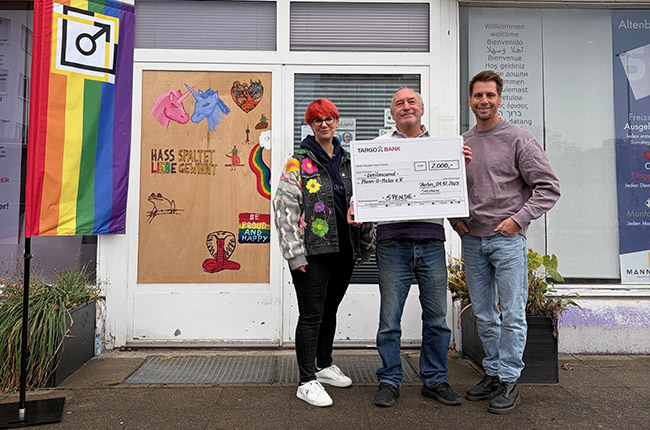 Drei Personen stehen vor einem Geschäft. Eine Person hält einen großen Scheck, auf dem "2000€" steht. Im Hintergrund sind eine bunte Flagge und ein Kunstwerk mit Einhörnern und dem Text "HASS SPALTET, LIEBE EINIGT" sichtbar. Die Personen tragen lässige Kleidung und es sind Pflanzen in Töpfen vor dem Geschäft.