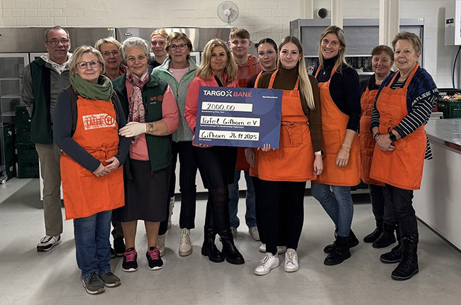 Eine Gruppe von Menschen steht in einer Küche, alle tragen orangefarbene Schürzen. In der Mitte hält eine Person einen großen Scheck von TARGOBANK über 2000,00 Euro für den Verein Tafeln Lichten e.V. Die Umgebung ist hell und modern, mit Küchenausstattung im Hintergrund. Das Datum auf dem Scheck ist der 26.11.2023.