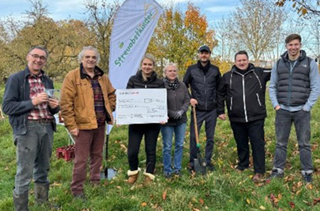 Eine Gruppe von sieben Personen steht im Freien auf einer Wiese, umgeben von Bäumen mit Herbstlaub. Sie halten einen großen Scheck und haben Spaten dabei. Im Hintergrund ist ein Fahne mit dem Aufdruck 'Streuobstklinik' sichtbar.