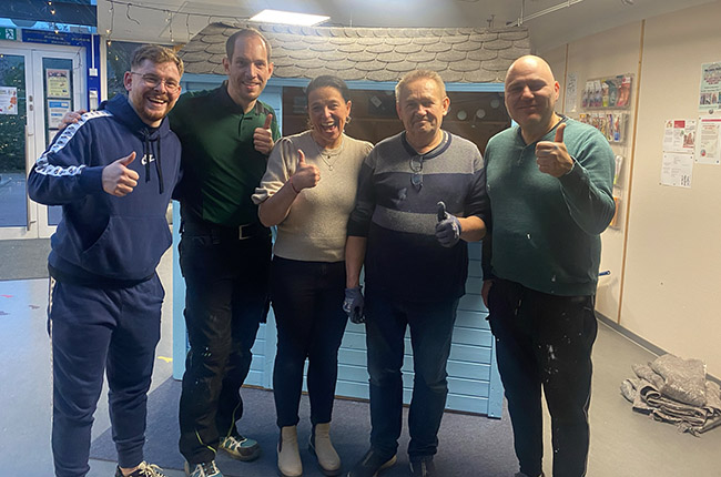 Fünf Personen stehen in einem Raum und posieren mit Daumen hoch. Sie sind in lässiger Kleidung und lächeln. Im Hintergrund ist eine blaue Wand mit einer Dachstruktur zu sehen.