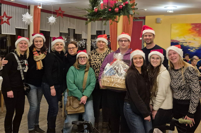 Eine Gruppe von Menschen steht zusammen in einem festlich dekorierten Raum. Alle tragen Weihnachtsmützen. In der Mitte hält jemand einen großen Geschenkkorb. Im Hintergrund sind Weihnachtsdekorationen und Teilnehmer der Feier zu sehen.