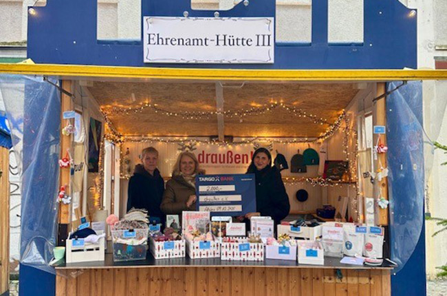 Ein Verkaufsstand namens 'Ehrenamt-Hütte III' mit Lichtern und Dekoration. Im Inneren stehen zwei Personen vor einem Tisch mit verschiedenen Produkten, darunter Karten und kleine Geschenke.