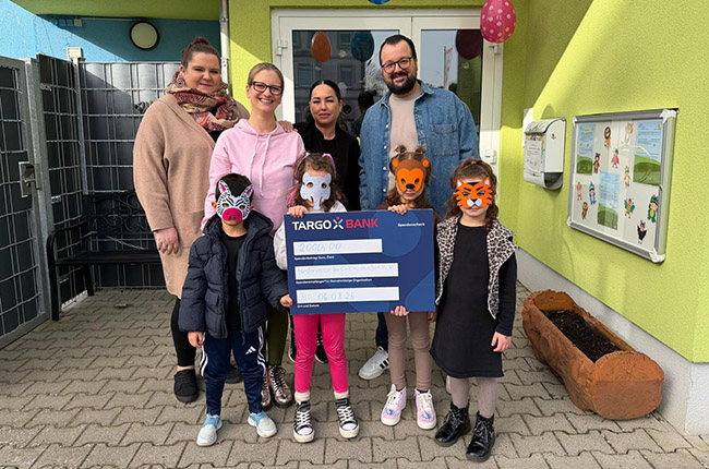 Eine Gruppe von sieben Personen steht vor einem Gebäude mit bunten Luftballons. Sie halten einen großen Scheck von TARGOBANK über 20.000,00 Euro. Die Kinder tragen Tiermasken, während die Erwachsenen lächeln. Im Hintergrund sind Fenster und eine Wand mit Zeichnungen sichtbar.