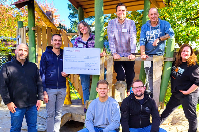 Eine Gruppe von sieben Personen steht und sitzt auf einem Spielplatz. Sie halten einen großen Scheck der TARGOBANK, während bunte Bäume im Hintergrund sichtbar sind.