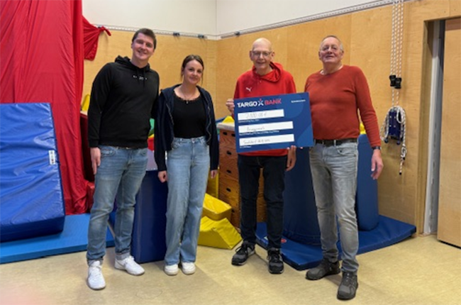 Vier Personen stehen in einem Raum mit Spielgeräten. Sie halten ein großes Scheck-Symbol der TARGOBANK. Der Raum hat eine Holzwand und bunte Matten.