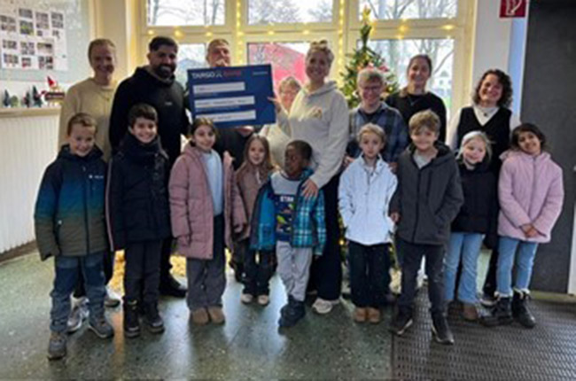 Eine Gruppe von Kindern steht zusammen in einem Raum mit großen Fenstern und einem Weihnachtsbaum im Hintergrund. Sie halten ein großes, blaues Schild mit dem Schriftzug 'TARGOBANK' und anderen Informationen. Die Kinder tragen verschiedene Winterjacken und sind fröhlich. Einige Erwachsene stehen hinter ihnen.
