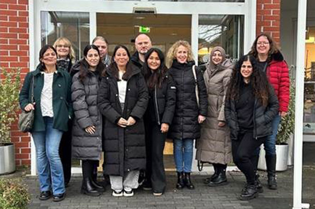 Eine Gruppe von zehn Frauen steht vor einem Gebäude mit großen Fenstern. Sie tragen lange Wintermäntel in verschiedenen Farben, darunter Schwarz, Grau und Rot. Die Frauen lächeln und posieren in einer lockeren Formation. Im Hintergrund sieht man eine Pflanze in einem Topf und die Eingangstür des Gebäudes. Das Wetter scheint kühl zu sein, was durch die Winterkleidung der Frauen angezeigt wird.