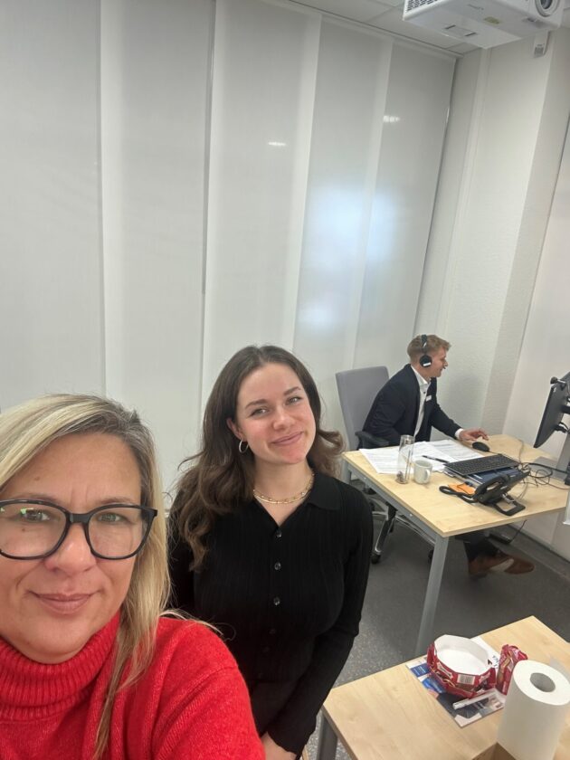 Ein Selfie von zwei Frauen in einem modernen Büro. Eine Frau trägt einen roten Pullover und lächelt, während die andere Frau mit langen, lockigen Haaren in einem schwarzen Oberteil im Hintergrund steht. Im Hintergrund ist ein Mann in einem Anzug zu sehen, der an einem Schreibtisch sitzt und mit einem Computer arbeitet. Auf dem Tisch sind verschiedene Gegenstände wie eine Tasse, Dokumente und eine Rolle Küchenpapier sichtbar.