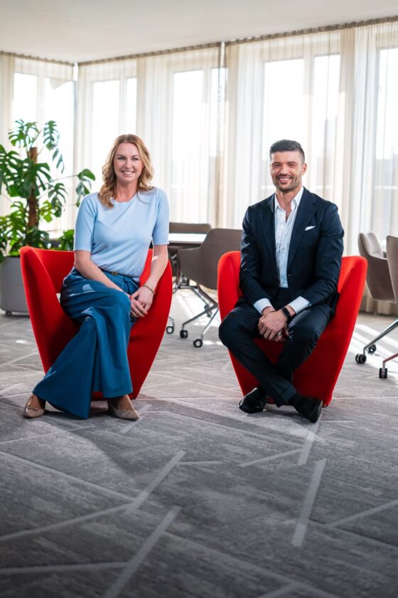 Eine Frau in einem hellblauen Oberteil und blauen weiten Hosen sitzt auf einem roten Stuhl neben einem Mann in einem schwarzen Anzug. Der Hintergrund zeigt große Fenster mit Vorhängen und einen modernen Raum mit Pflanzen und Möbeln.