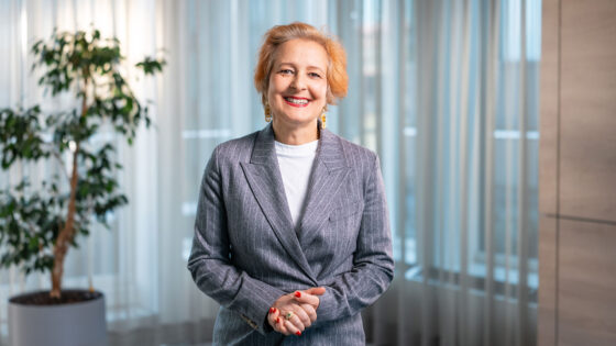 Eine Person steht in einem modernen Raum mit großen Fenstern und Vorhängen. Sie trägt einen grauen Blazer mit einem weißen T-Shirt darunter und hat die Hände vor sich gefaltet. Im Hintergrund ist ein großes, grünes Pflanzenarrangement in einem Topf sichtbar.