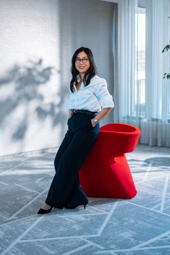 Eine Person steht lässig an einem roten Stuhl in einem modernen Raum. Sie trägt ein weißes Hemd und schwarze, weite Hosen. Der Raum hat große Fenster, durch die Tageslicht strömt, und einen hellen Teppich mit geometrischem Muster.