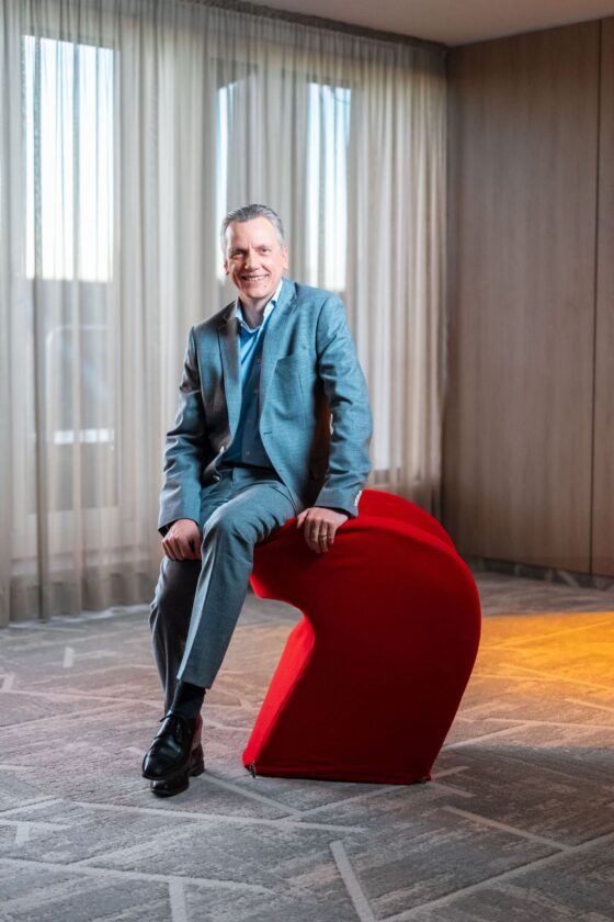 Eine Person sitzt auf einem roten, modernen Stuhl in einem Raum mit großen Fenstern, die mit hellen Vorhängen bedeckt sind. Die Person trägt einen grauen Anzug und ein blaues Hemd.