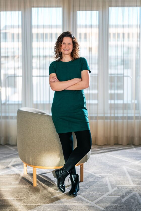 Eine Person steht mit verschränkten Armen in einem Raum mit großen Fenstern. Sie trägt ein grünes Kleid und schwarze Strumpfhosen sowie Stiefel. Im Hintergrund ist ein runder, grauer Sessel zu sehen.