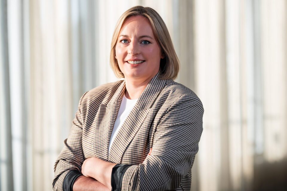 Eine Person mit schulterlangen, blonden Haaren steht mit übergeschlagenen Armen. Sie trägt einen karierten Blazer über einem weißen Oberteil. Der Hintergrund besteht aus sanft drapierten Vorhängen, die ein weiches Licht hereinlassen.